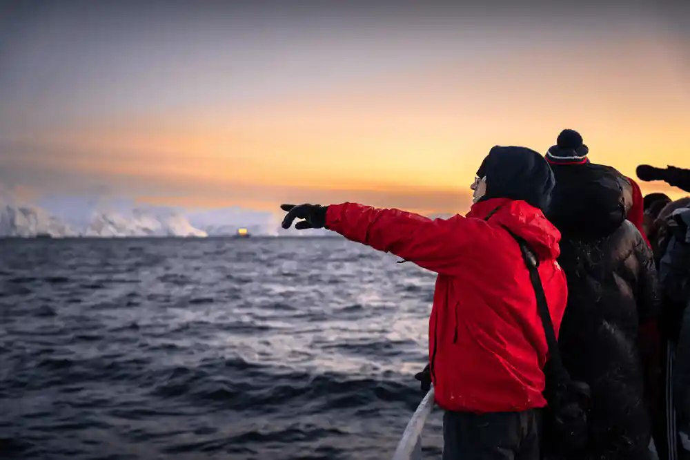 A passenger in a red jacket pointing out across the water at sunset