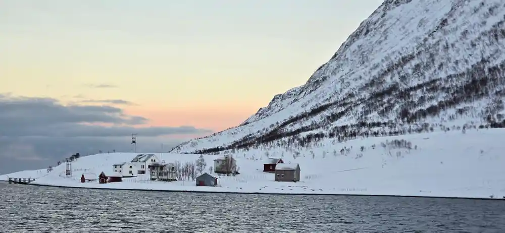 Red-painted farm buildings under a steep snow mountain at the shore
